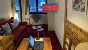 A cozy room with a red sofa, a wooden table with a laptop, a TV on the wall, and a window with a view of mountains. - Cosy 2-room apartment in the heart of Chamonix, Mont Blanc view (Chamonix-Mont-Blanc)