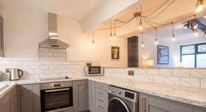 A modern kitchen with a built-in oven, microwave, and washing machine, marble countertops, and pendant lighting.
