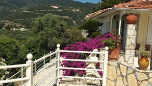 Eine steinerne Treppe führt zu einem Haus mit Terrakotta-Dachziegeln, umgeben von üppigem Grün und Bergen im Hintergrund.