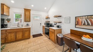 A kitchen with wooden cabinets, a sink, a stove, and a dining area with a table and chairs. - GrandLake*Kayak*Forest View*Firepit*King*Lakefront (Grove)