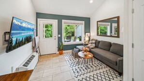A living room with a gray sofa, a coffee table with flowers, a large TV, and a view of greenery outside. - GrandLake*Kayak*Forest View*Firepit*King*Lakefront (Grove)