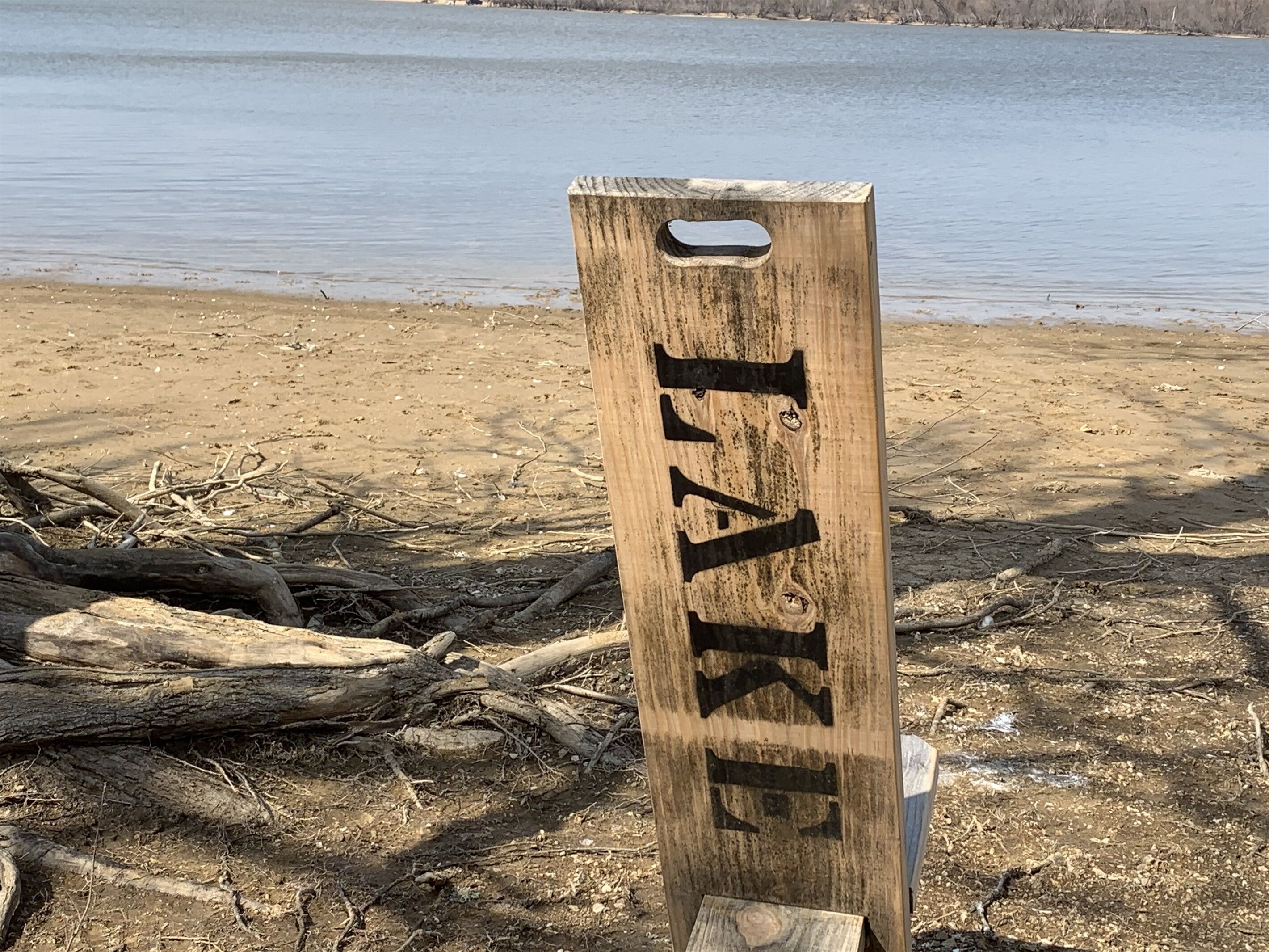 Een houten bord met de tekst 'TAKE' op een strand met drijfhout en een meer op de achtergrond.