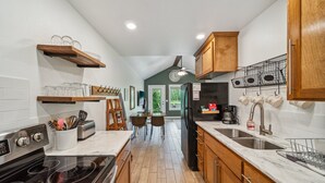 A kitchen with a stove, oven, sink, and wooden cabinets. There are hanging mugs and a clock on the wall.