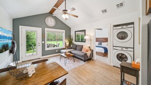 A cozy living room with a sofa, coffee table, and a dining area with a wooden table and chairs. There is a ceiling fan, a clock, and a washing machine in the background.