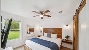 A bedroom with a bed, a ceiling fan, a TV, a window, and a wooden headboard.