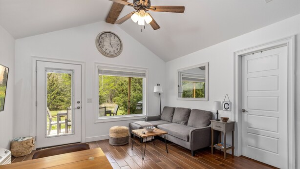 Un salon spacieux avec un canapé gris, une table basse en bois et un ventilateur de plafond. De grandes fenêtres offrent une vue sur l’extérieur.