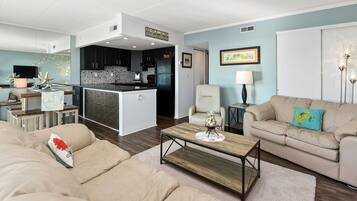 A modern living room with a beige sofa, a wooden coffee table, and a kitchen area with dark cabinets and a black refrigerator.