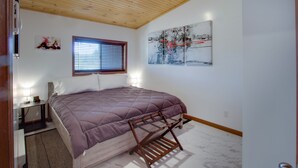 Un dormitorio con techo de madera, una cama con colcha púrpura, una ventana con persianas, un cuadro abstracto colgado en la pared y una mesita de noche con lámpara.