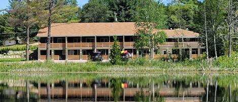 A two-story building with a flat roof, surrounded by trees and a body of water.