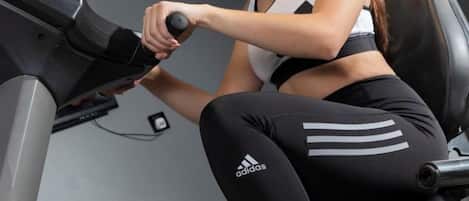 Una persona usando una bicicleta estática, vestida con mallas negras con un logotipo blanco, en un gimnasio.
