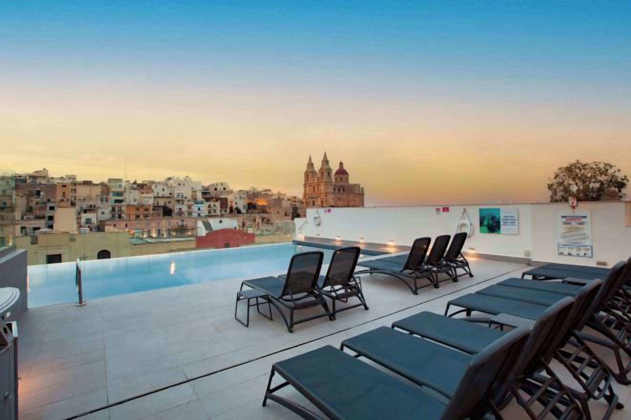 A rooftop pool area with lounge chairs and a view of a cityscape at sunset.