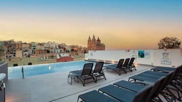 A rooftop pool area with lounge chairs and a view of a cityscape at sunset.
