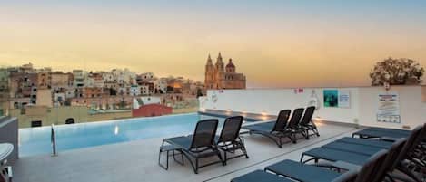 A rooftop pool area with lounge chairs and a view of a cityscape at sunset.