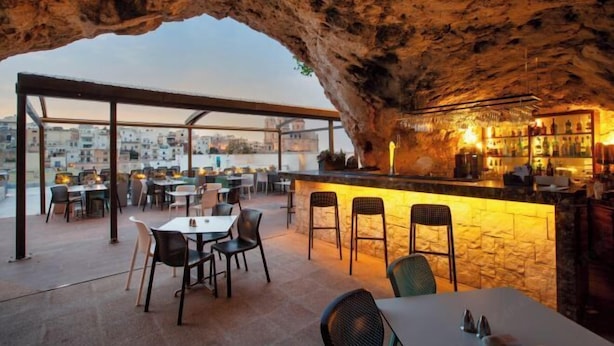 An outdoor dining area with a stone wall and a bar, featuring tables and chairs, and a view of the cityscape. - Pergola Hotel & Spa (Mellieha)