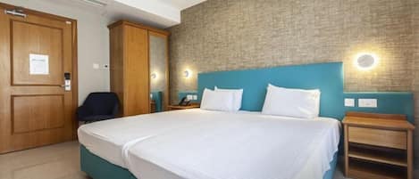 A hotel room with a large bed, a wooden wardrobe, a desk with a chair, and a wall-mounted light fixture.