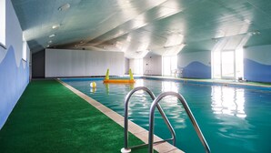 An indoor swimming pool with a green artificial turf border, a metal pool ladder, and a clear ceiling with skylights. - Stone Cottage Retreat with Pool & Lakeside (St Clears)