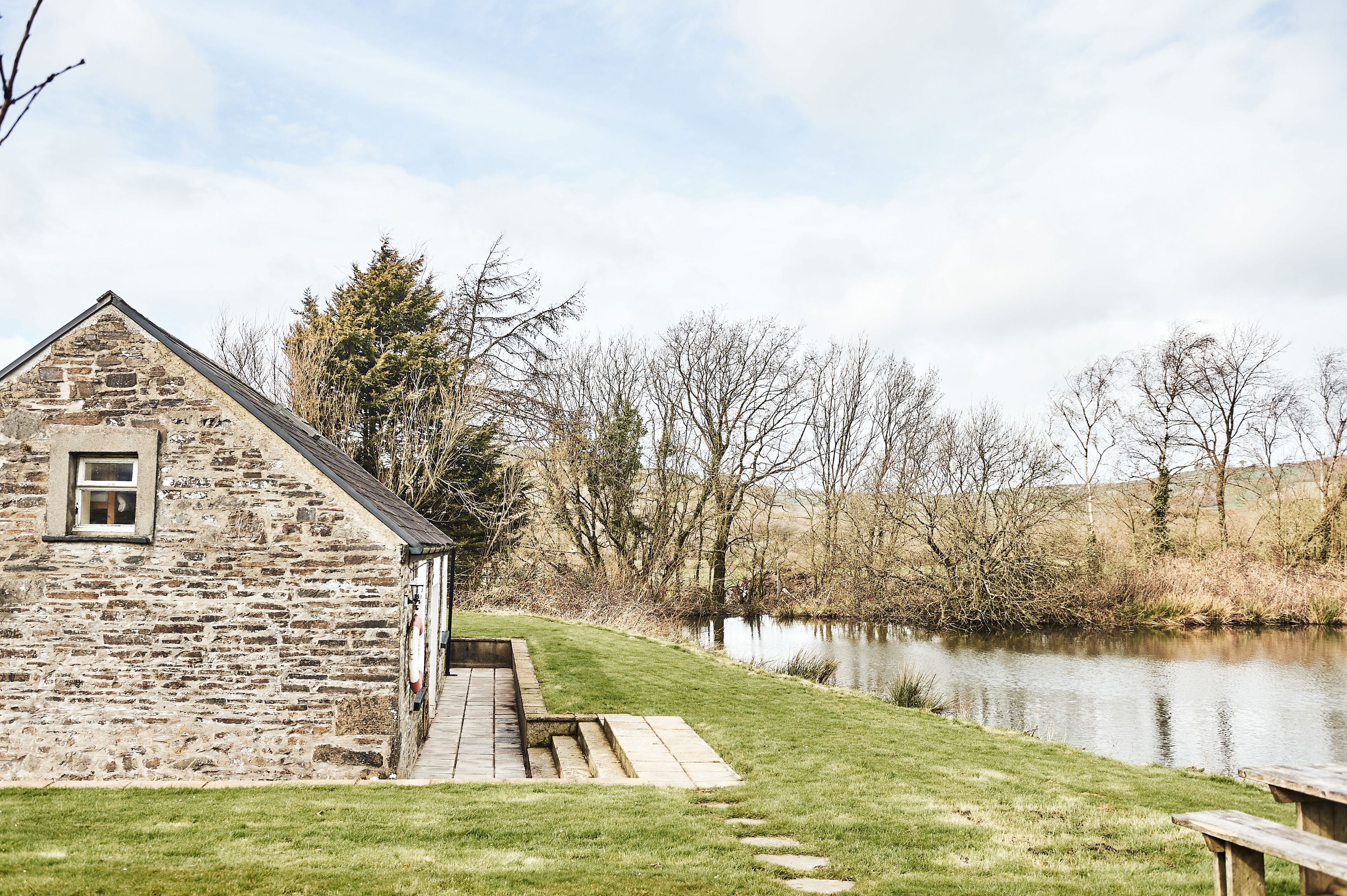 A stone building with a small window and a wooden deck by a calm lake, surrounded by trees and grassy areas.