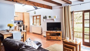 A cozy living room with a leather sofa, wooden coffee table, and a television. A kitchen is visible in the background with wooden cabinets and a window. - Stone Cottage Retreat with Pool & Lakeside (St Clears)
