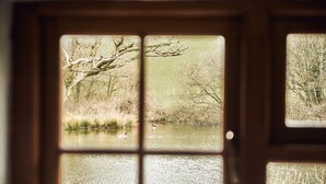 A view through a window with a wooden frame, displaying a calm body of water, trees, and reeds. - Stone Cottage Retreat with Pool & Lakeside (St Clears)