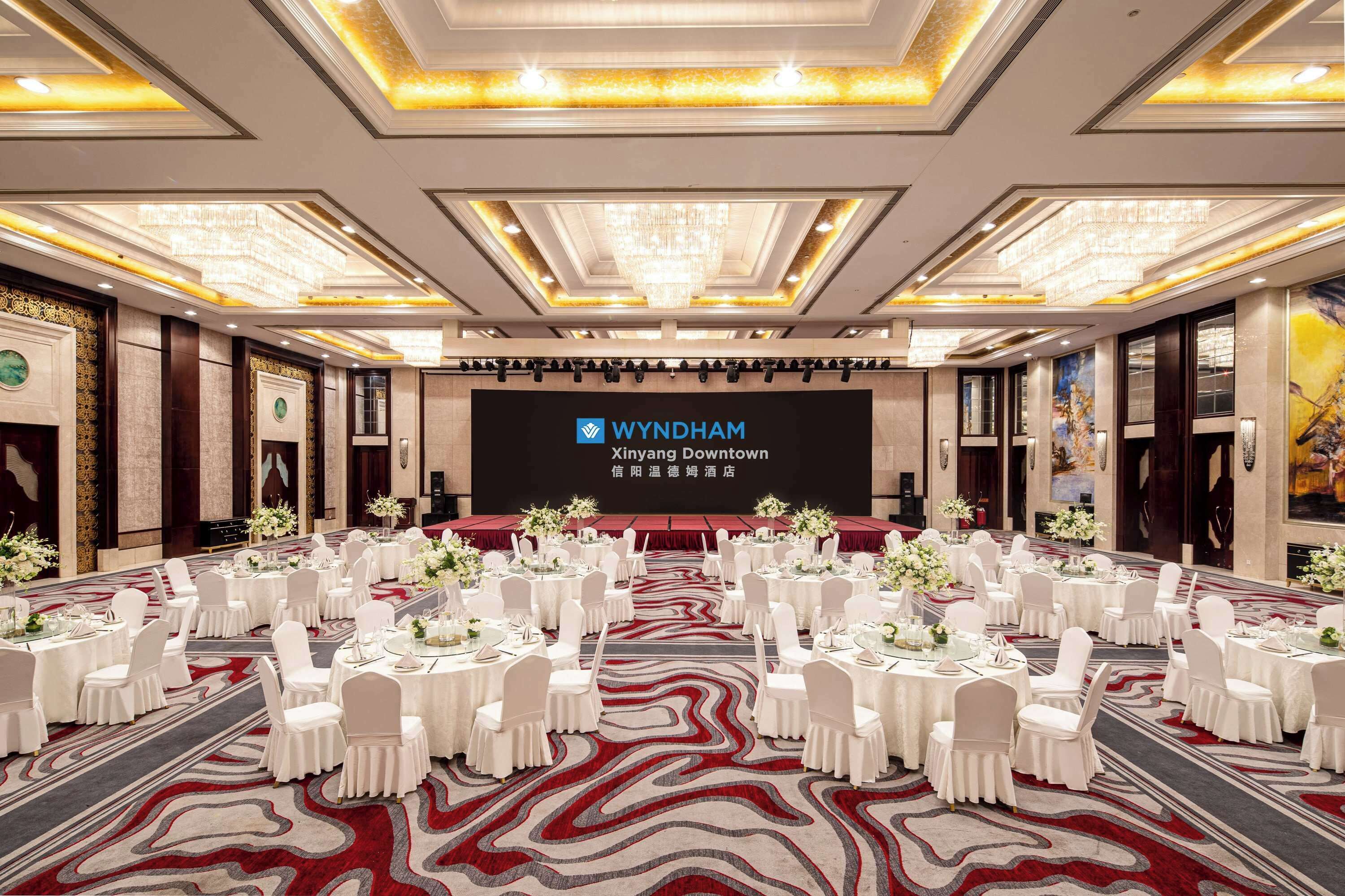 A conference room at Wyndham Xinyang Downtown with round tables, white chairs, and a large screen displaying the hotel's name.