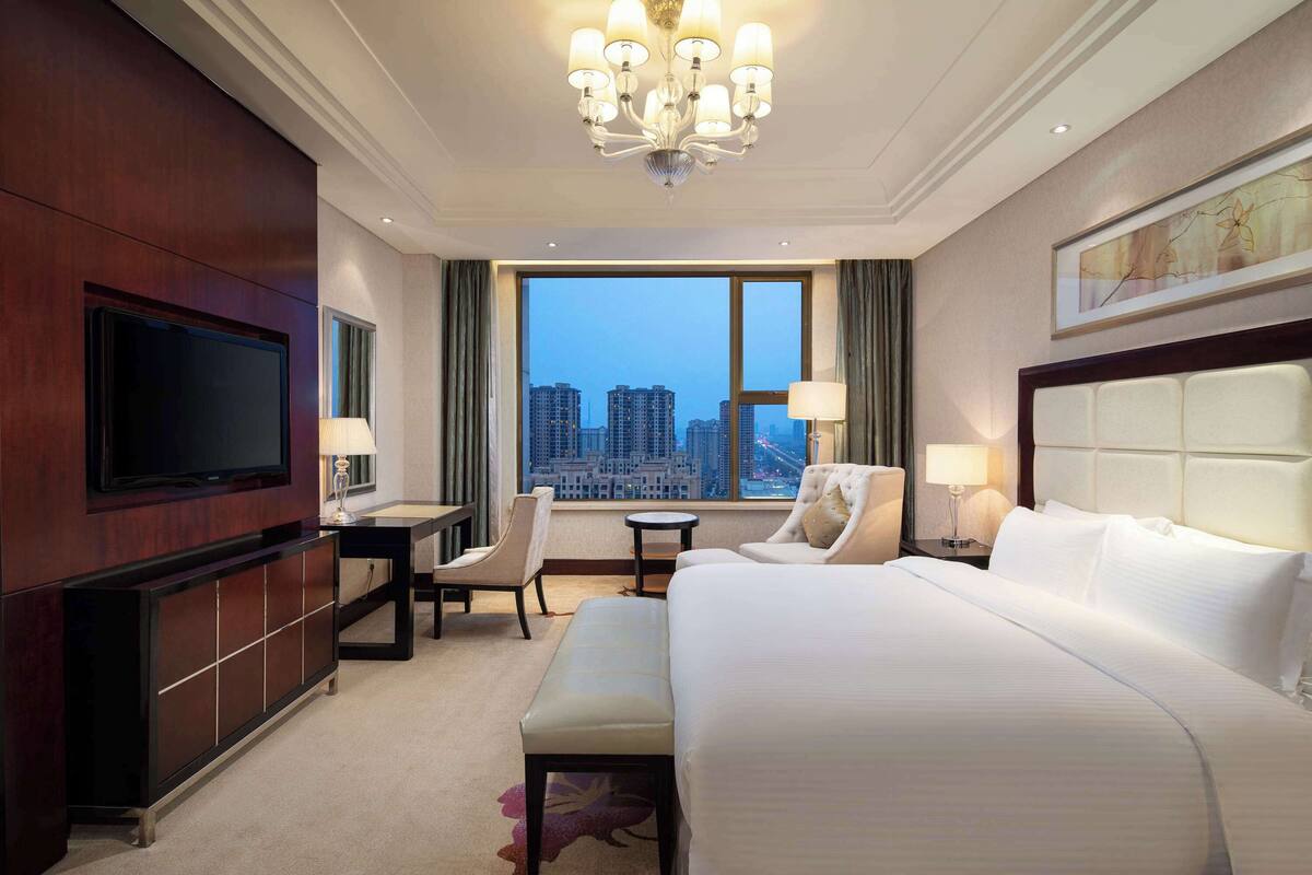 A hotel room with a large bed, a television, a desk with a chair, a small table, and a view of the cityscape through the window.