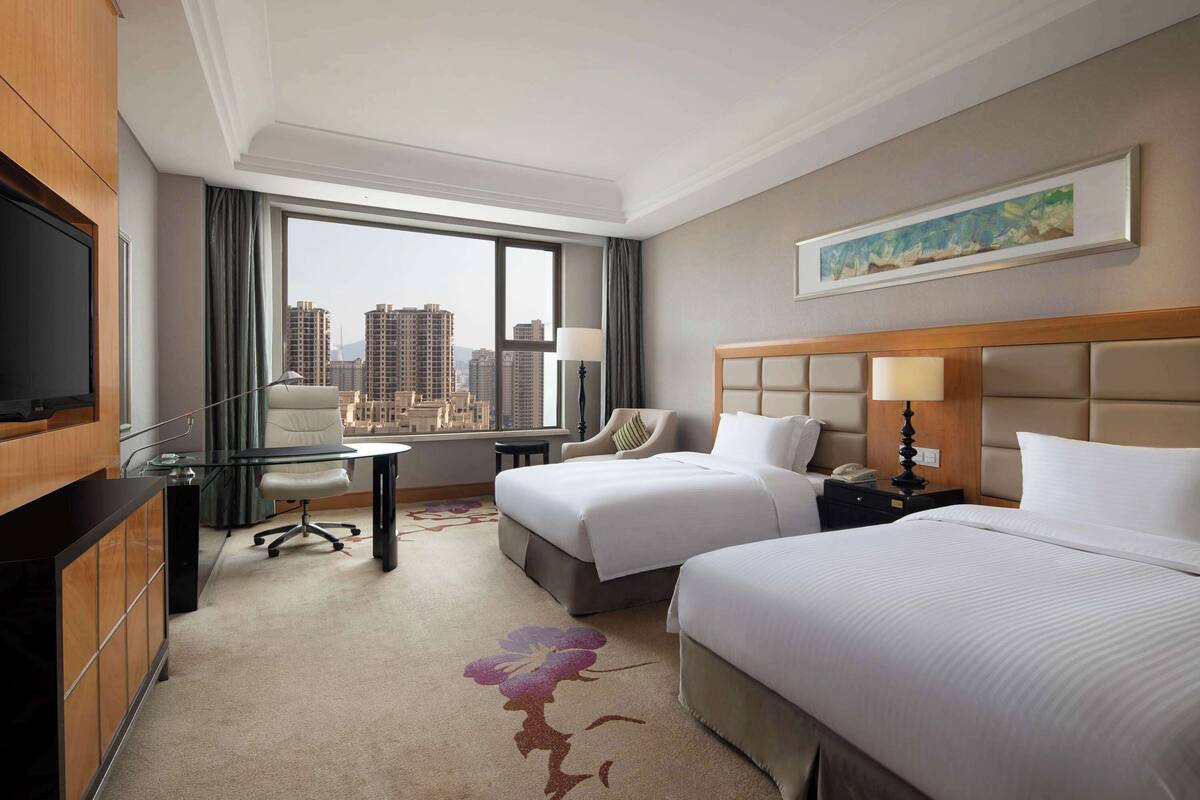 A hotel room with two beds, a desk, a TV, and a large window with a city view.