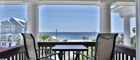 A balcony with two chairs and a table, overlooking a beach and ocean.