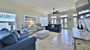 A spacious living room with a blue sofa, a patterned armchair, and a blue couch. There is a ceiling fan, a flat-screen TV, and a wooden coffee table. - ECDI | PCB Near 30-A 5BR 4BA Pool Hot Tub EV Charger Early Booking Discounts (Panama City Beach)