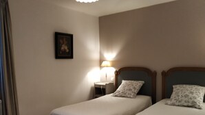 Habitación de hotel con dos camas individuales, una mesita de noche con lámpara, una silla pequeña y un cuadro en la pared.