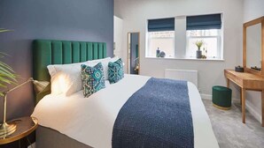 Een moderne slaapkamer met een bekleed hoofdeinde in groen, een witte spreidek en kussens met een patroon. Er staat een nachtkastje met een lamp, een raam met uitzicht, en een houten dressoir.
