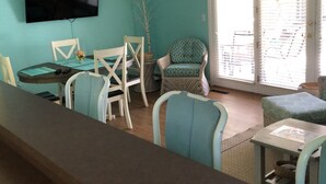 Una sala de estar con una televisión montada en la pared, una mesa de comedor con sillas, un sofá y una ventana con persianas.