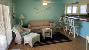 Una sala de estar acogedora con un sofá, dos sillones de mimbre, una mesita y una zona de cocina con barra y taburetes.