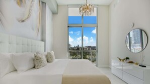 A modern bedroom with a large bed, a white dresser, a round mirror, and a city view through large windows. - One Bedroom Condo King Bed With City Views (Miami)
