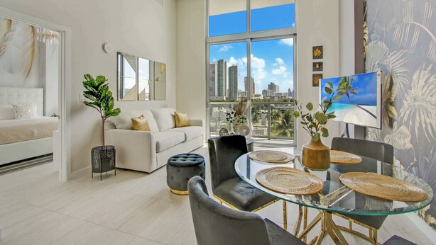 A modern living room with a glass dining table, comfortable seating, a large window with a city view, and a decorative plant. - One Bedroom Condo King Bed With City Views (Miami)