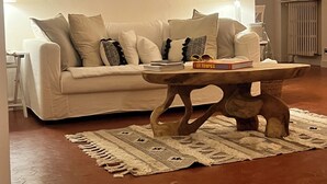 Uma sala de estar com um sofá bege, um espelho redondo, uma mesa de centro com livros e um tapete estampado.
