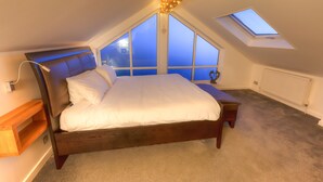 A bedroom with a sloped ceiling, a skylight, a bed with white bedding, a bedside table, and a wooden shelf. - Modern & Cosy Home with Hot-Tub, 5min drive to Beach, ideal for large families. (Goldsithney)