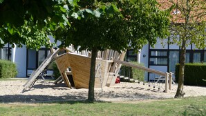 A playground with a wooden slide and swings, surrounded by trees and a building in the background.