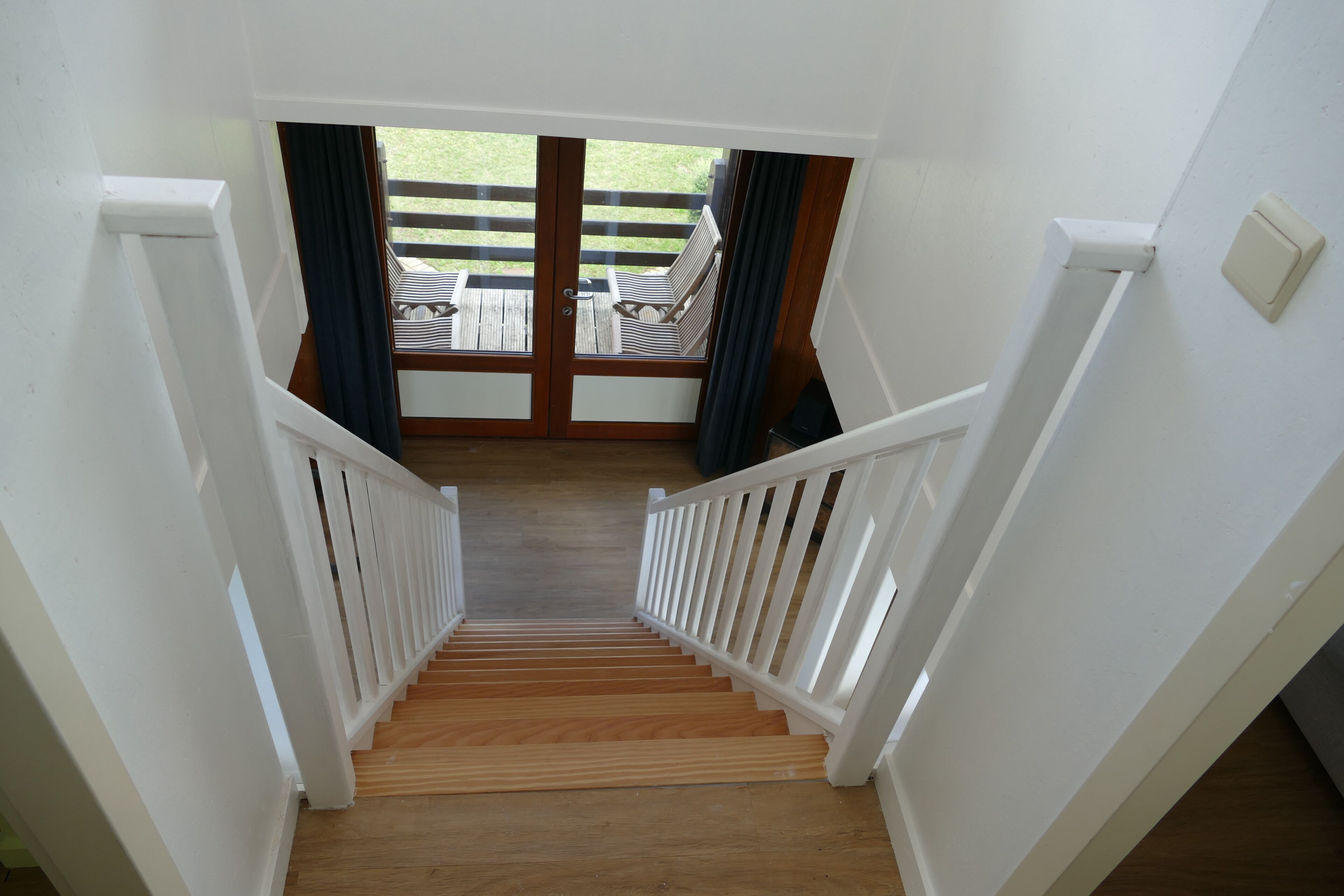 A wooden staircase with white railings leading to a balcony with a view of a grassy area and outdoor furniture.