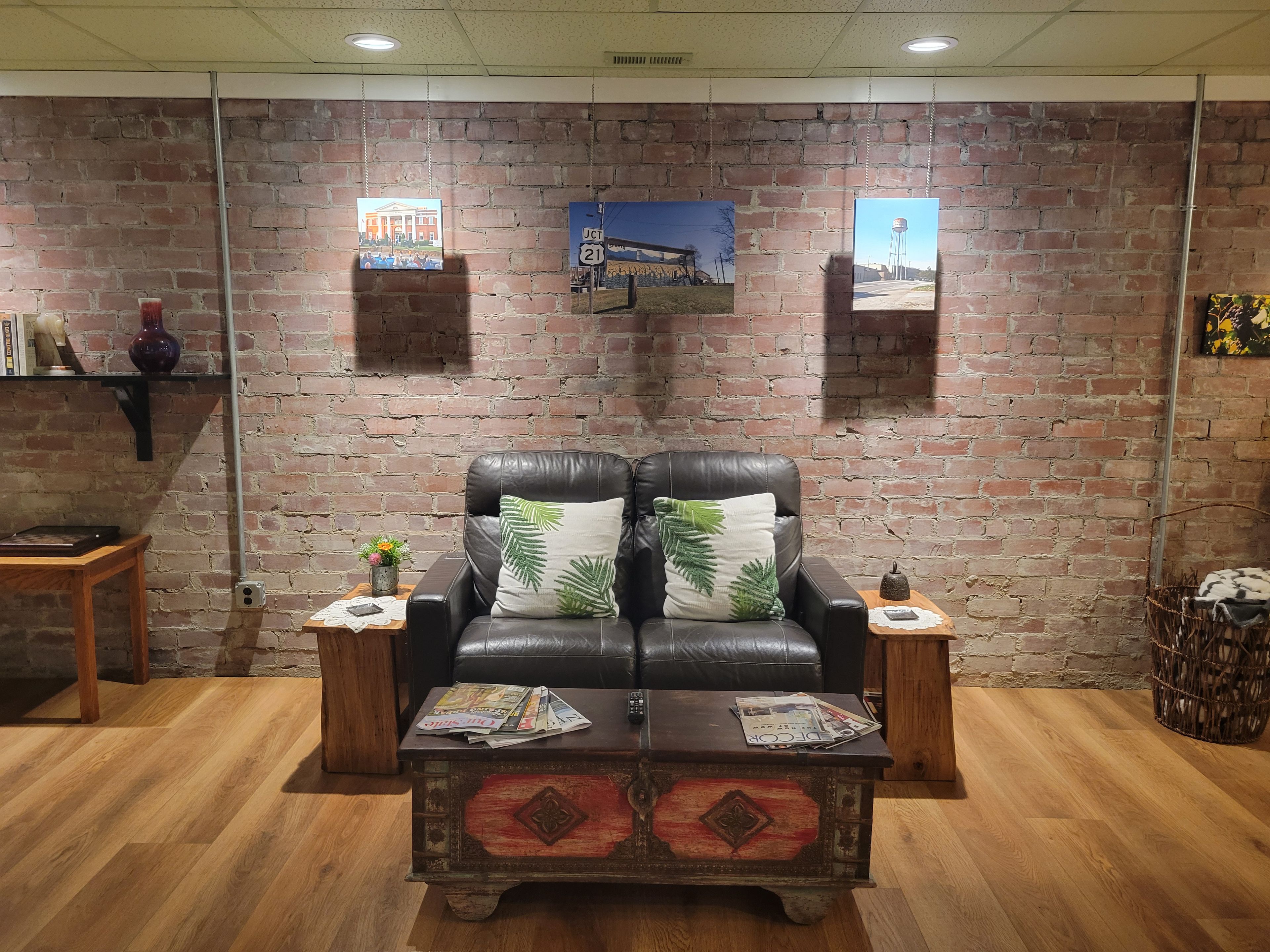 A room with a brick wall, a leather sofa with green leaf cushions, a wooden coffee table with magazines, and a small side table with a vase and a plant.