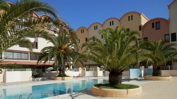 A swimming pool area with palm trees and a building with a curved roof in the background. - La Brigantine - appartement avec piscine proche plage (Gruissan)