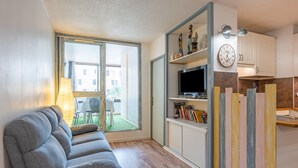 A modern living room with a grey sofa, a TV on a shelf, and a large window overlooking a green outdoor area. - La Brigantine - appartement avec piscine proche plage (Gruissan)
