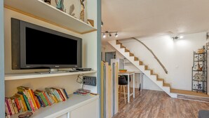 A modern living room with a flat-screen TV, bookshelves, a staircase, and a dining area. - La Brigantine - appartement avec piscine proche plage (Gruissan)