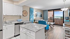A modern kitchen with white cabinets, a built-in dishwasher, and a marble countertop. There is a dining area with a blue chair and a table, and a large window offering a view of the ocean.