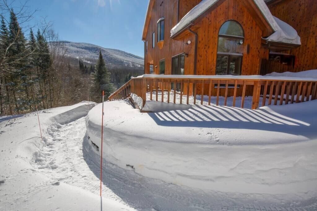 A wooden cabin with a snow-covered deck and a clear view of the surrounding forest and mountains.