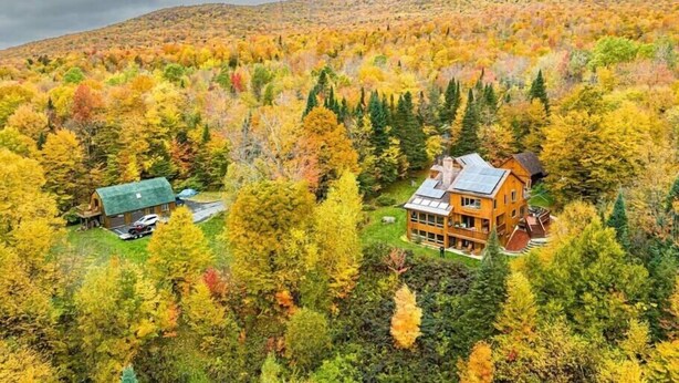 Una cabaña con un edificio central grande y una estructura más pequeña, rodeada de un bosque denso y montañas al fondo.