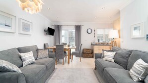 A modern living room with grey sofas, a dining table, and a TV mounted on the wall. - Sea Shells, St Ives – Sleeps 4 – On-Site Parking – Dog Friendly – Balcony (St Ives)