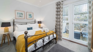 A neatly made bed with a metal frame, two bedside lamps, and a yellow throw blanket. There are framed pictures on the wall and a view of the outside balcony through a window with curtains. - Sea Shells, St Ives – Sleeps 4 – On-Site Parking – Dog Friendly – Balcony (St Ives)