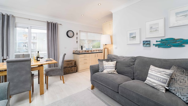 A modern living room with a grey sofa, dining table, and chairs. There are framed pictures on the wall and a lamp on the side table. - Sea Shells, St Ives – Sleeps 4 – On-Site Parking – Dog Friendly – Balcony (St Ives)