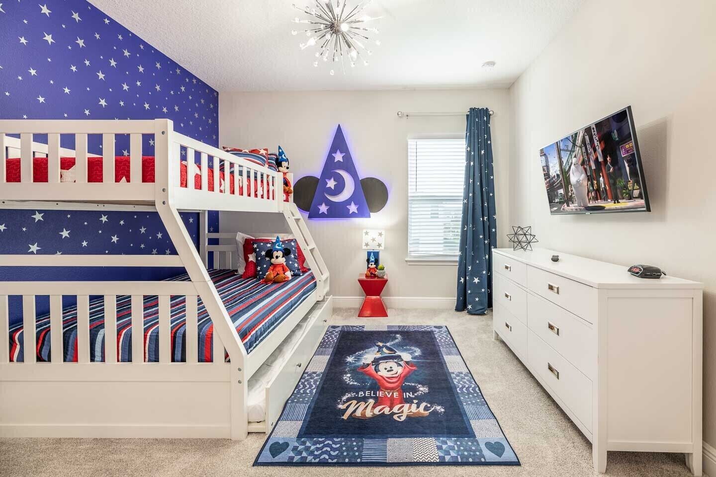 Une chambre d’enfant avec un lit superposé, un bureau, une commode, un tapis décoré aux motifs de Mickey Mouse et un élément décoratif mural.