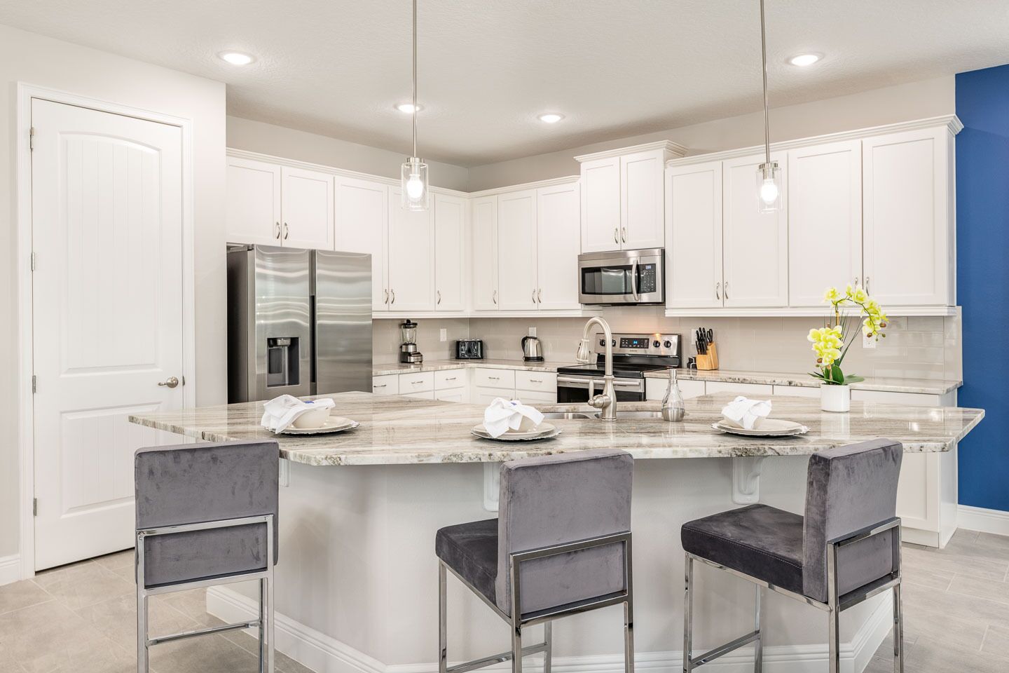 A modern kitchen with white cabinets, stainless steel appliances, a marble island with bar stools, and pendant lighting.